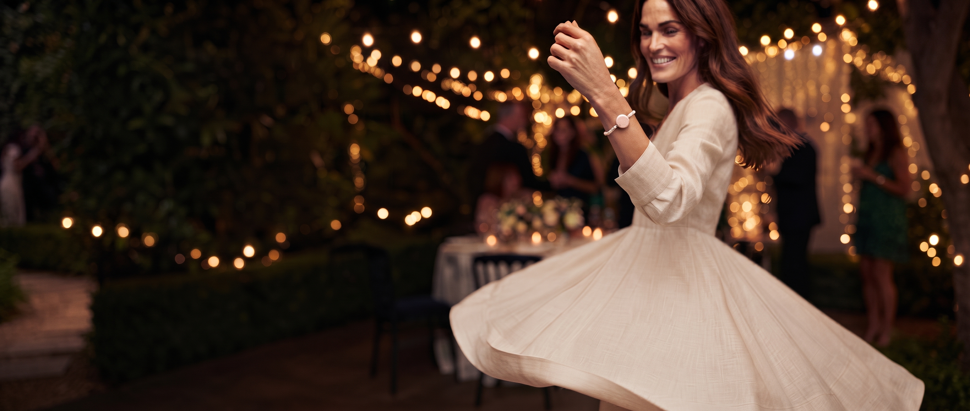 A woman in a cream linen dress dances at an evening garden party, her raised wrist wearing a Juni bracelet with a pale pink Charm